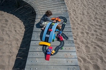 wobbel board posé sur des planches en bois, au dessus du sable, à la mer. Un circuit de voitures est posé sur la planche et autour. L'enfant joue avec le circuit