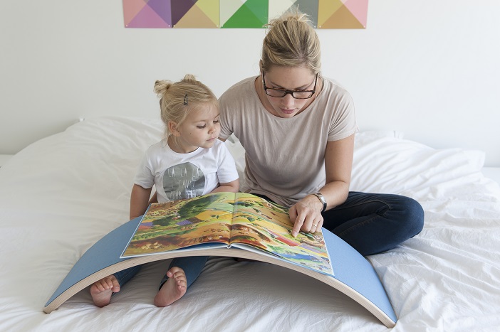 une maman et sa fille assises sur un lit. Une wobbel board bleue est posée au dessus de leur jambe, la maman porte des lunettes et lit un livre à sa fille. Le livre est posé sur la wobble board